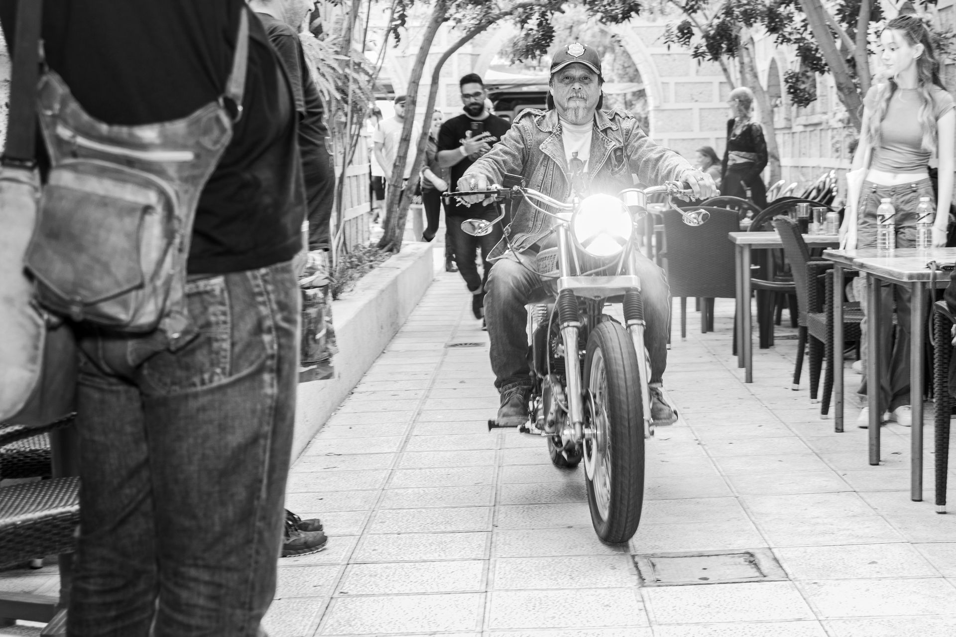 A man is riding a motorcycle down a sidewalk in a black and white photo.