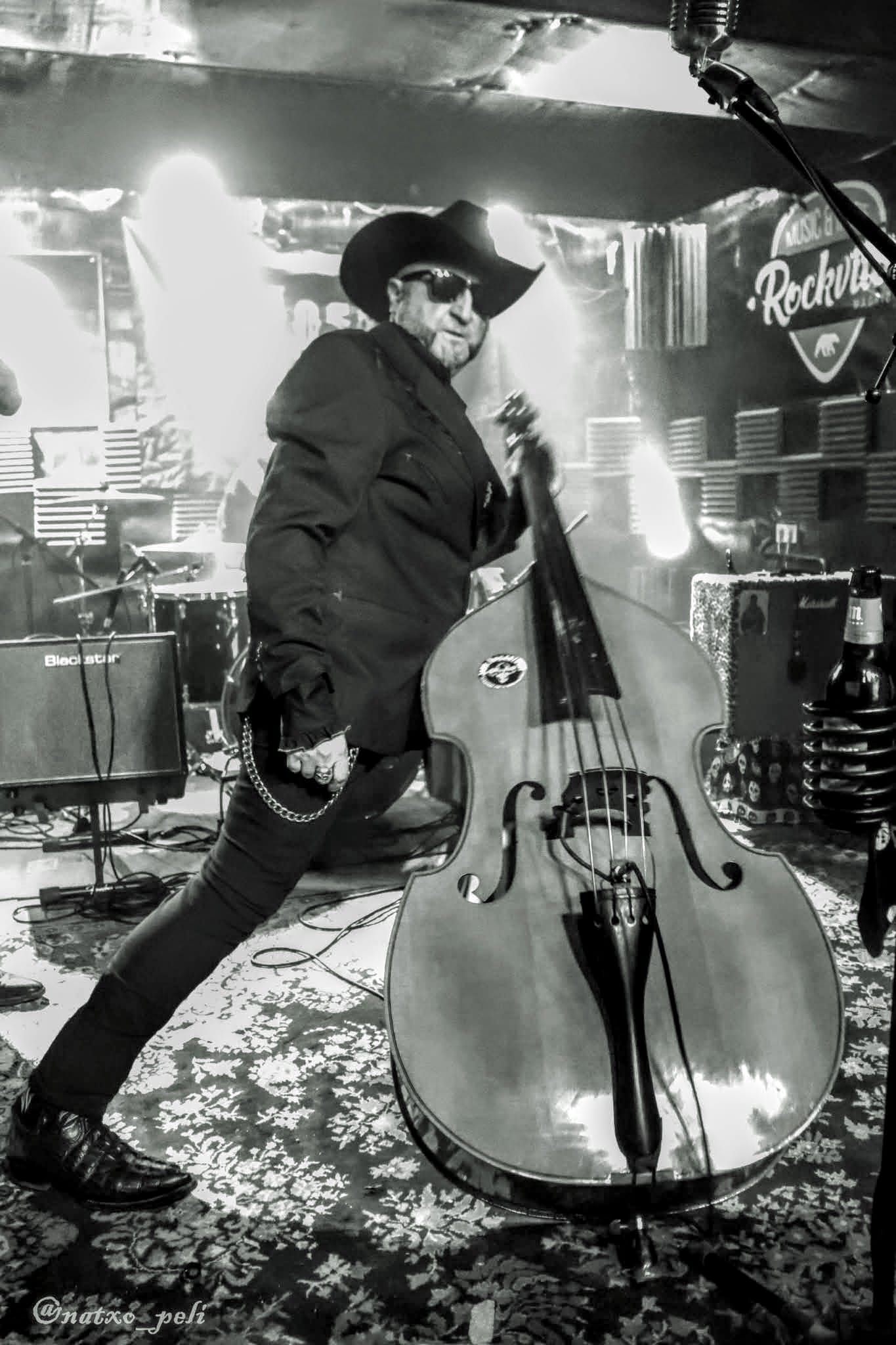 Two men on stage, one singing into a microphone, the other playing a stand-up bass. Red and blue stage lighting.
