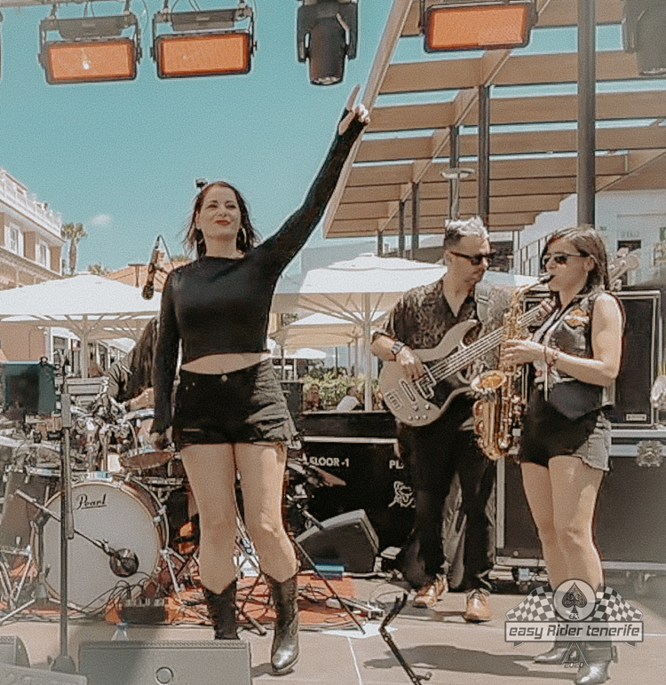 A woman in a black top is standing on a stage with a man playing a guitar and a woman playing saxophone