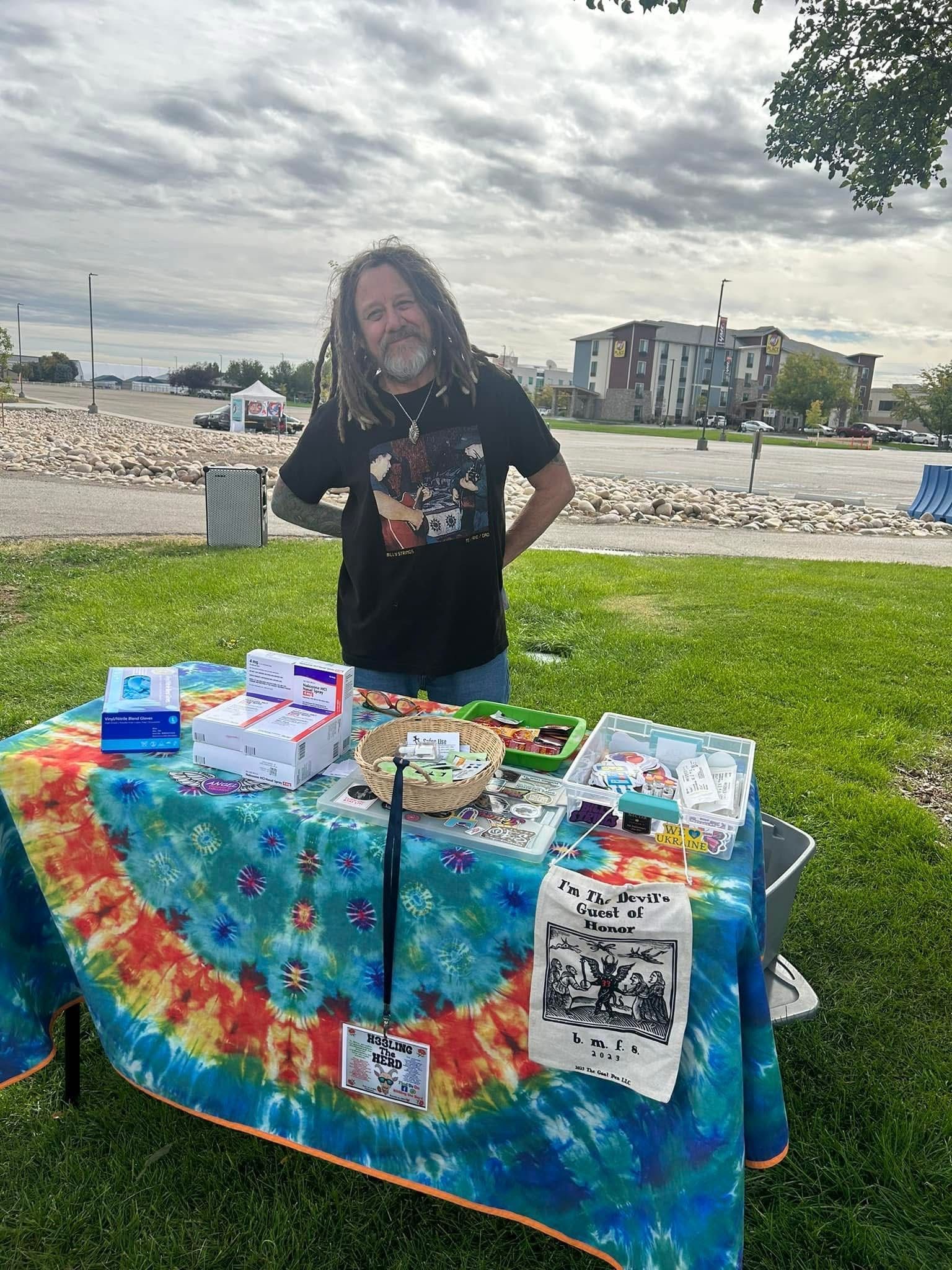 A man with dreadlocks is standing in front of a table in a park.
