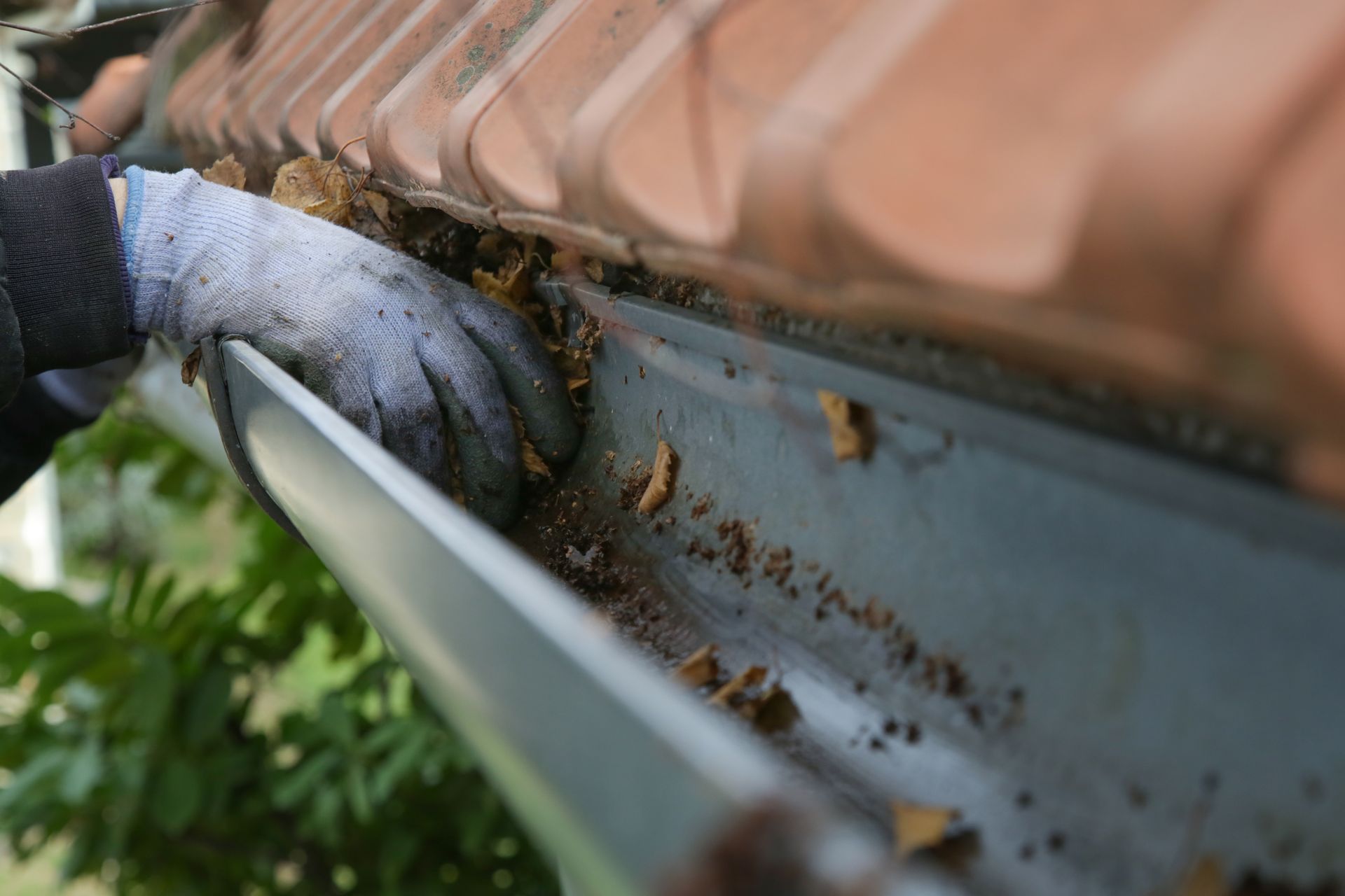 Gloved hand cleaning debris from a rain gutter beside a brick roof edge