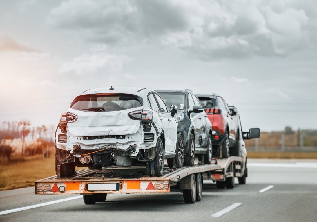Cars damaged in a crash loaded on a flatbed truck on a highway under cloudy skies.