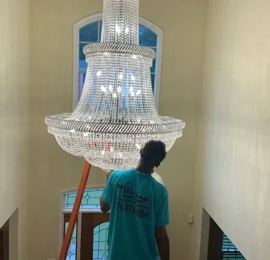 Person on ladder cleaning a large, ornate crystal chandelier in a foyer.