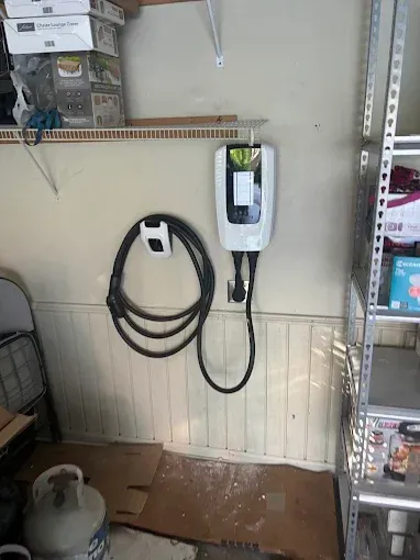 An electric vehicle charging station mounted on a garage wall. Black charging cable hangs.