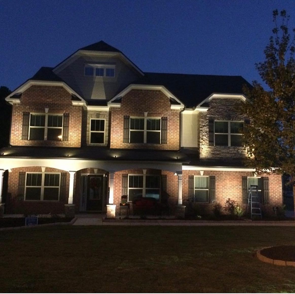 Illuminated two-story brick house at dusk, with porch, shutters, and spotlights.