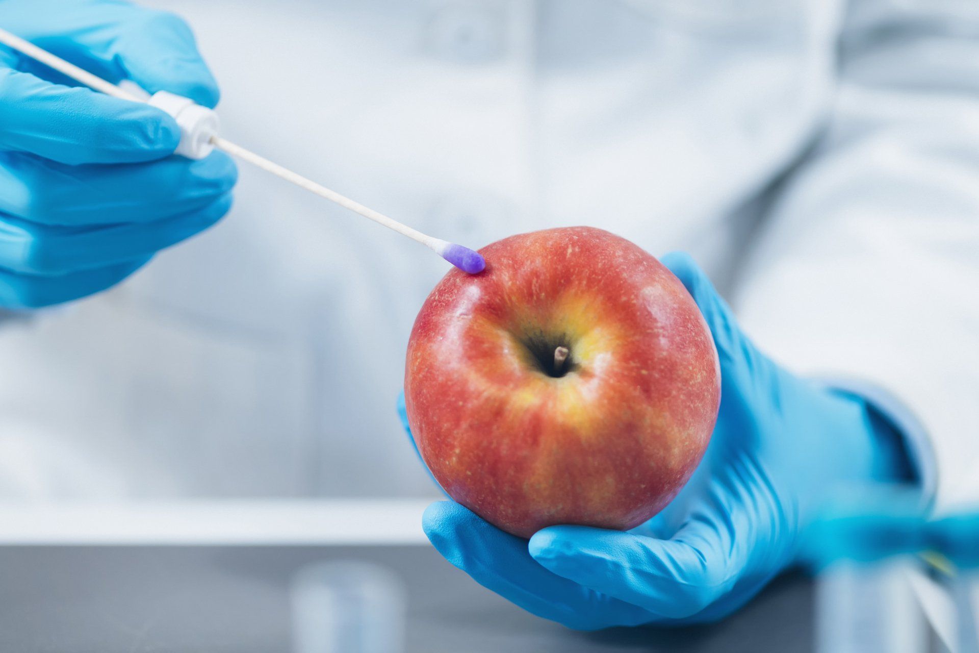 A person wearing blue gloves is holding an apple and a swab.