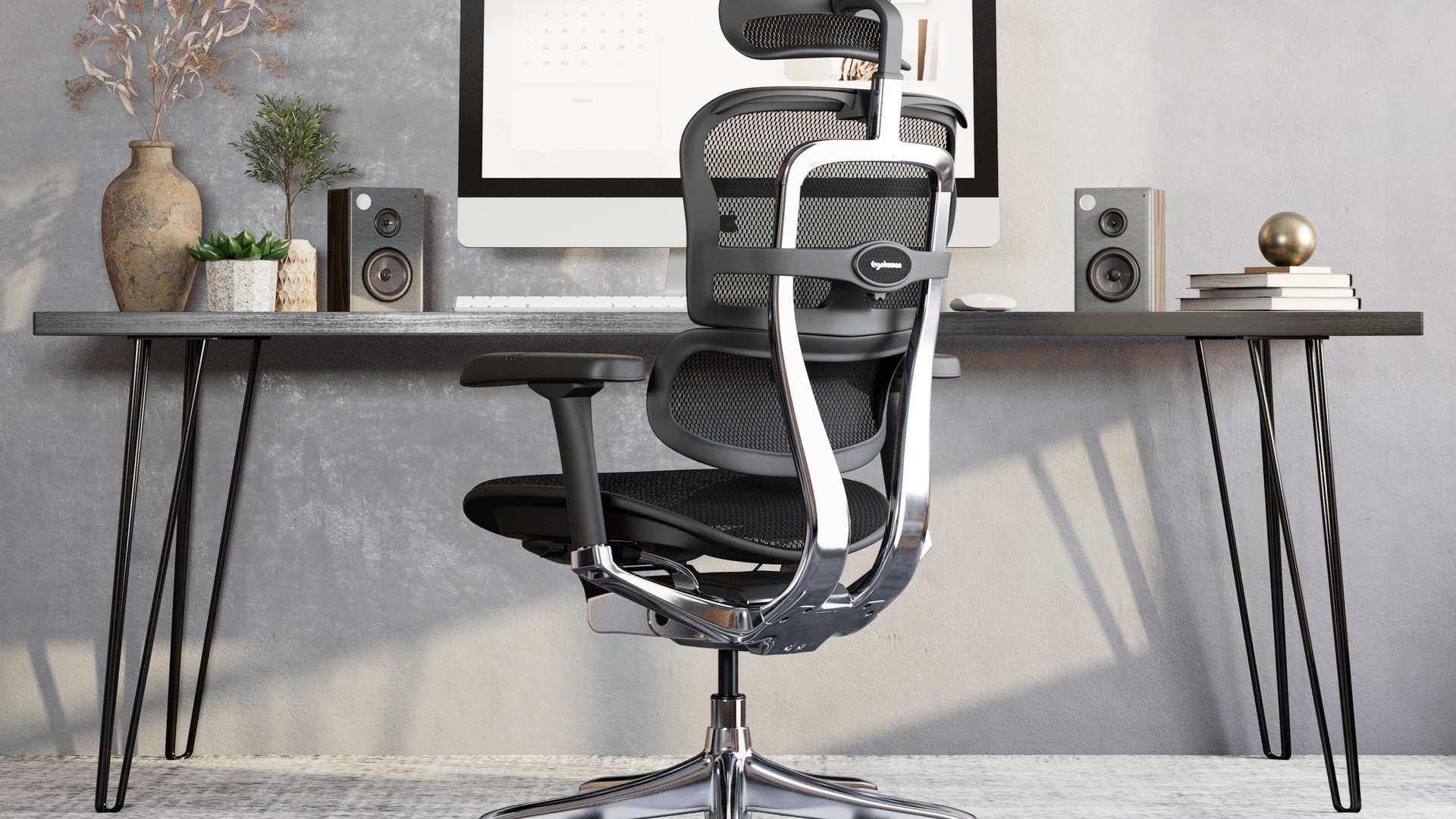 Black Ergohuman Elite office chair situated at a black desk with hairpin legs. The carpet and walls are grey. On the desk is an iMac, speakers, plants and books 