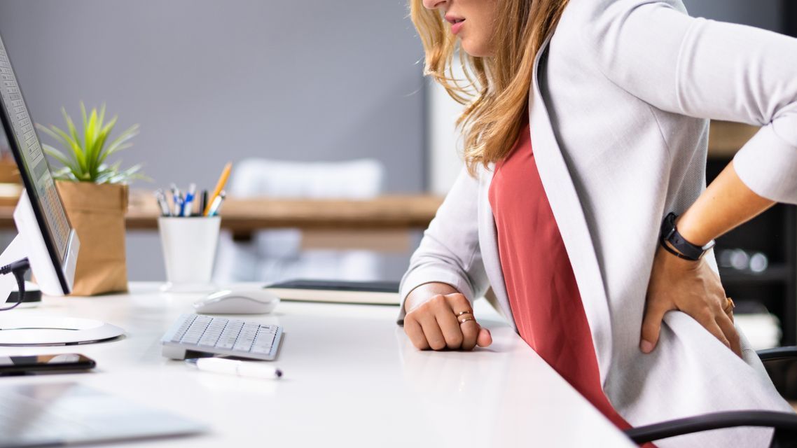 Woman working at a desk wearing a white blazer and pink top holding her back in pain as she sits. 