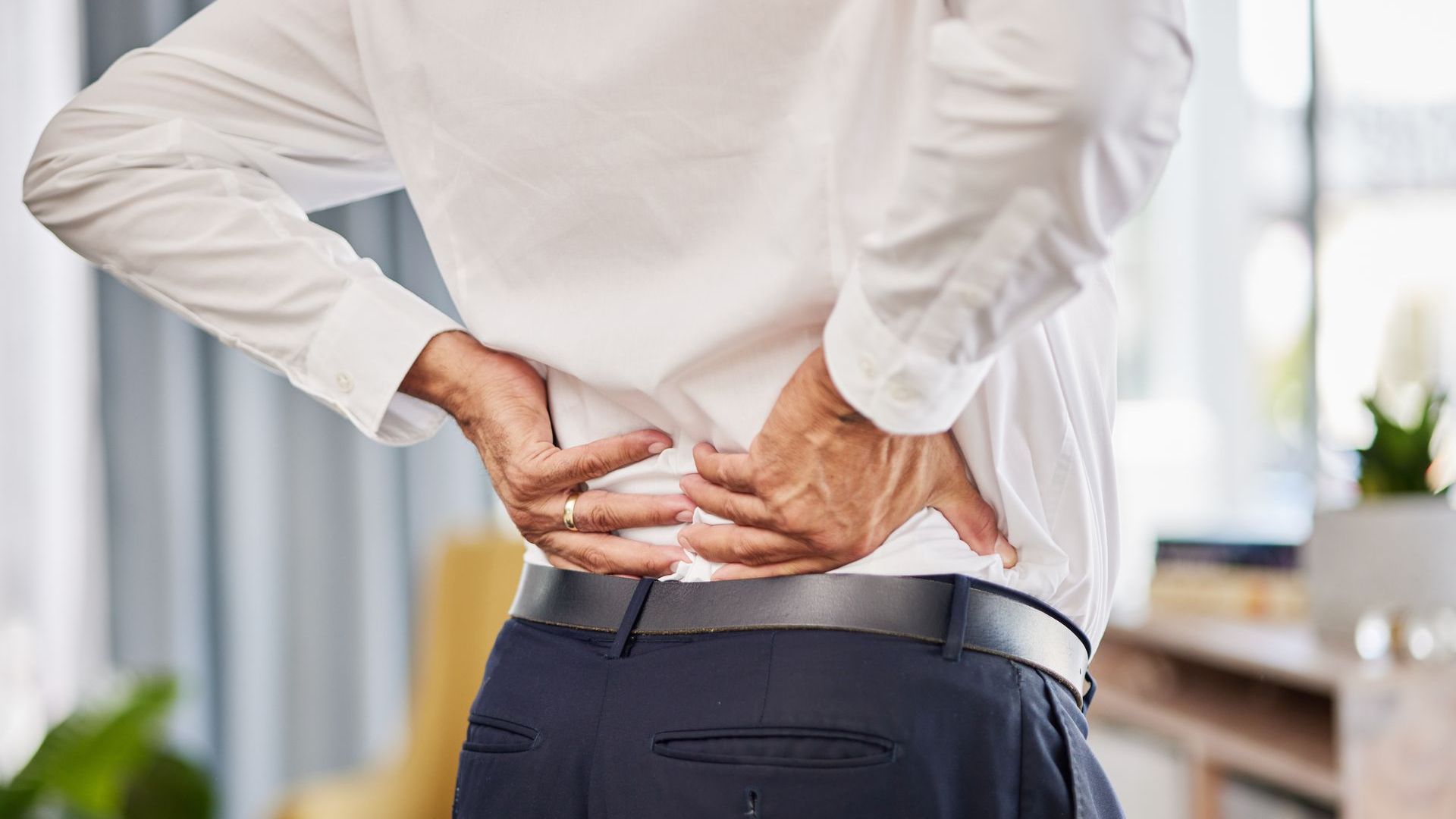 Close up image of a man's lower back. He is holding his back in pain. The man wears a white shirt and blue trousers with a black belt. He is wearing a wedding ring. 