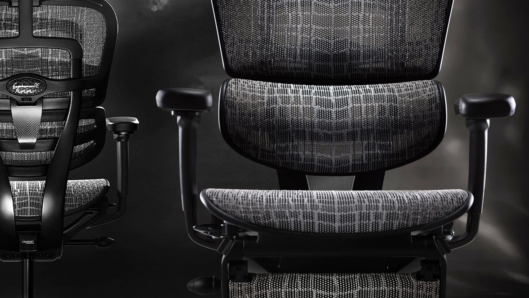 Two Ergohuman Carbon gaming chairs against a black smokey background. One chair is facing the camera, and the other is facing away. 