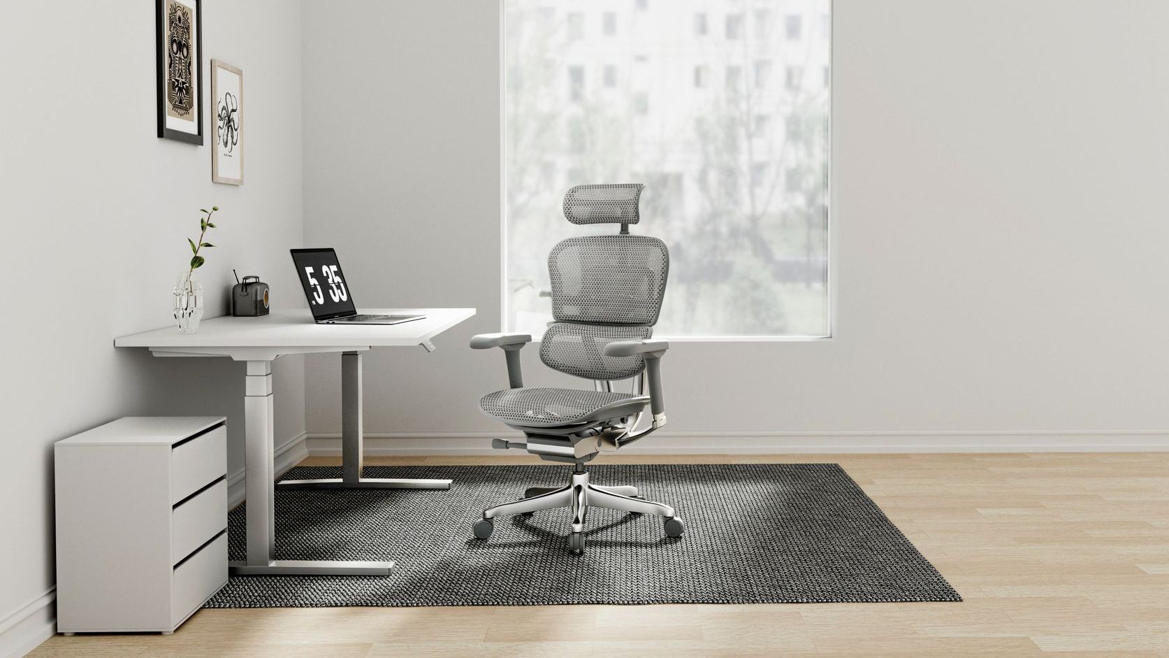 Grey Ergohuman chair on a black rug at a white desk looking at a laptop. There is a white wall and a soft window behind the chair. 