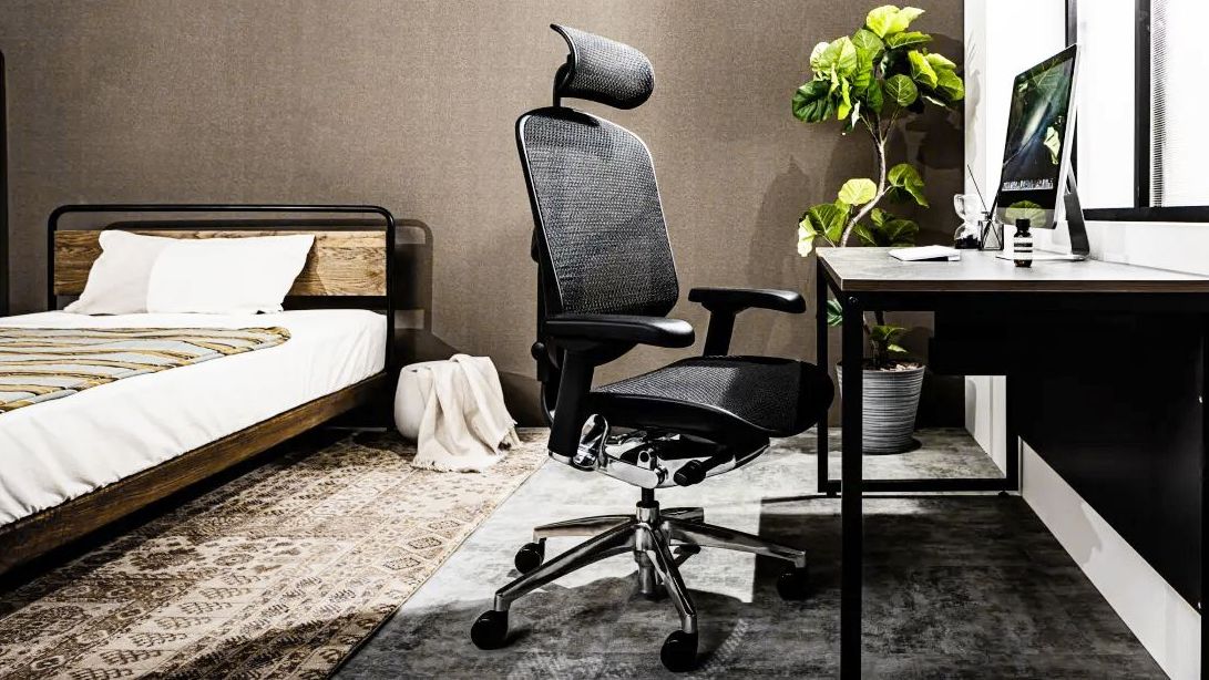 Black mesh office chair facing at a desk in a bedroom. On the desk there is a monitor and a small amber bottle. In the background is a healthy green plant. 