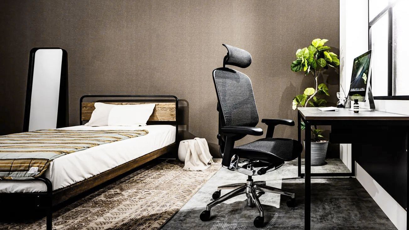 A black mesh office chair sits at a desk in a home office environment. A bed and mirror sit behind.