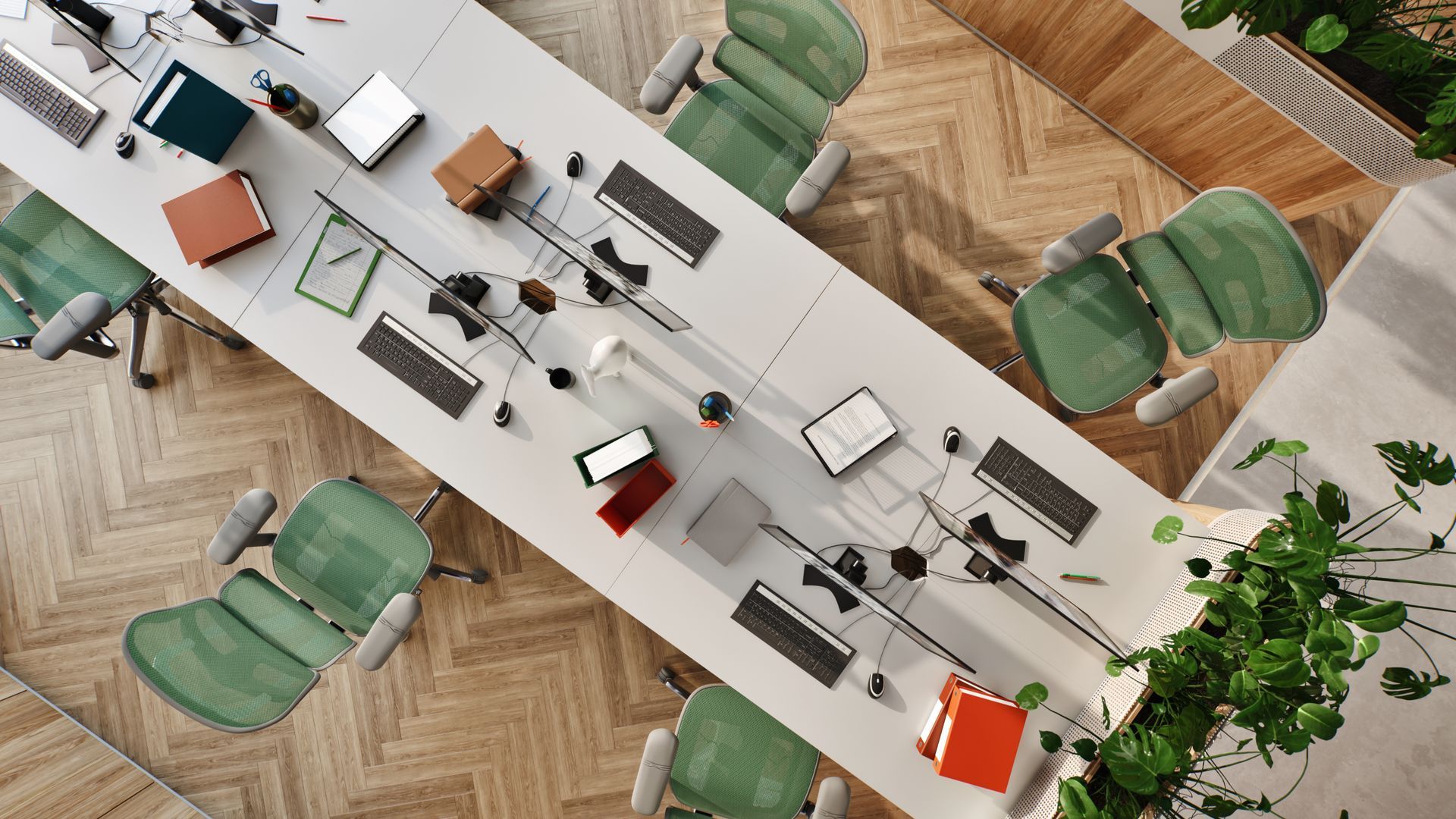 Birdseye view of an office with a white desk in the middle, with six chairs at each section of the large desk. The chairs are green mesh Ergohuman office chairs in a grey frame. At each desk is a computer, keyboard, notebooks, and some books. The floor is wooden herringbone. The office is adorned with green plants. 