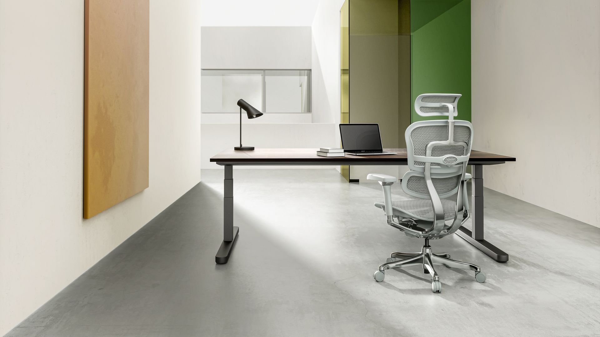 Grey on grey Ergohuman Elite chair in an open office space, situated at a desk. On the desk is a laptop, two books and a black lamp. 