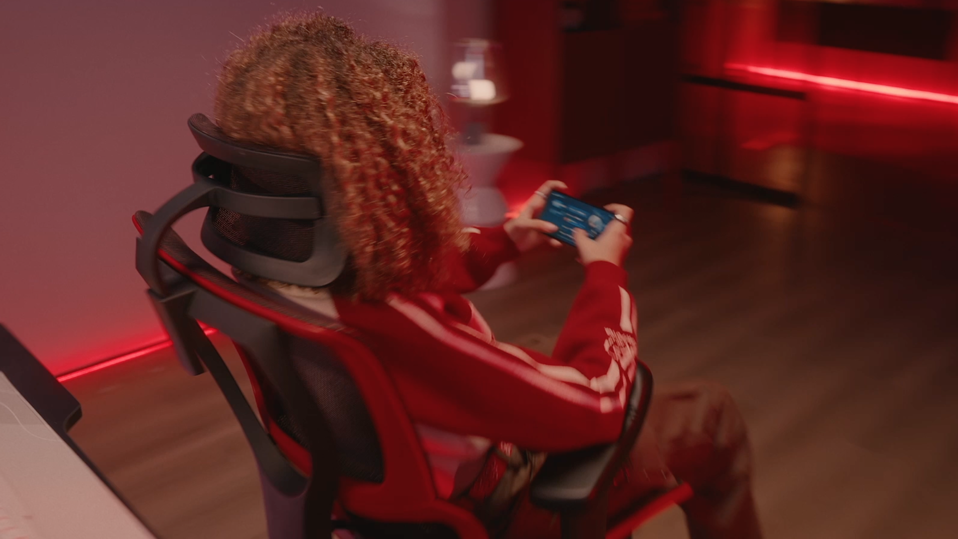 Back top right view of a girl with copper curly hair playing a smartphone game while sitting in a red Mirus Ultra gaming chair. 