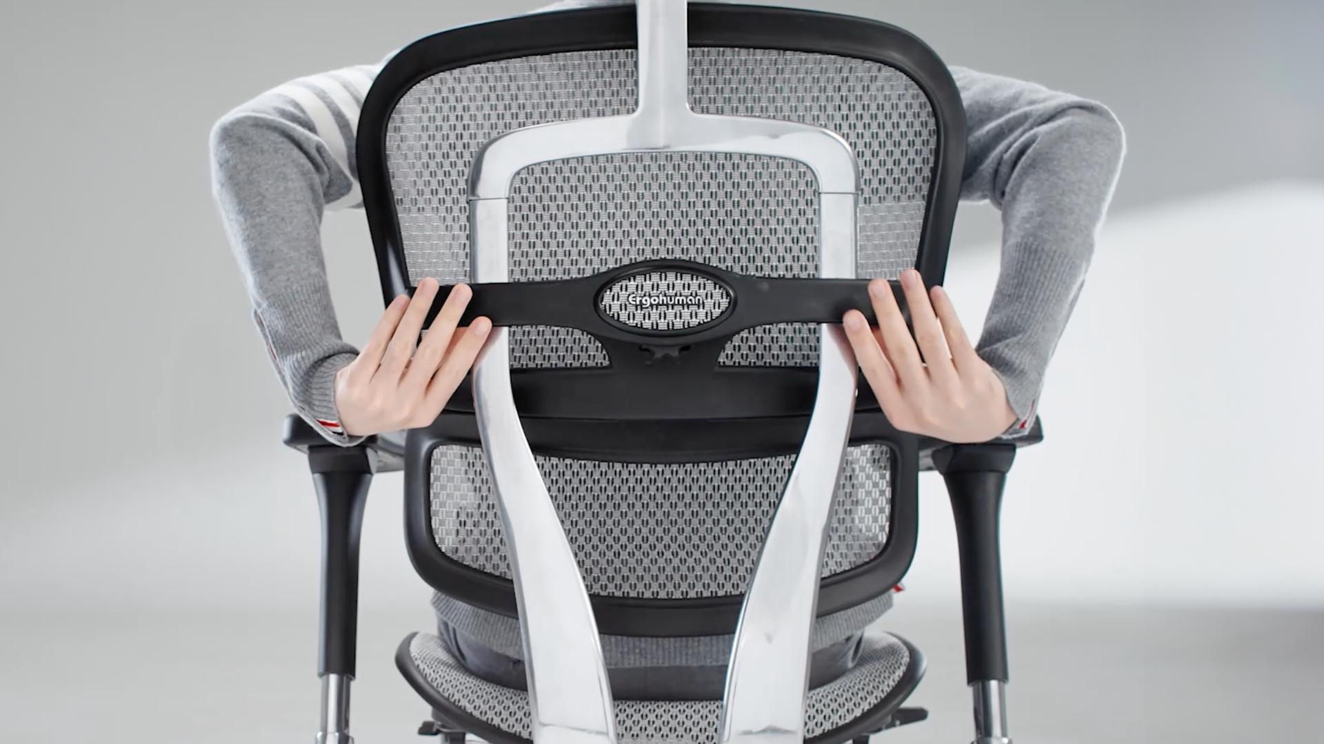 The back of the Ergohuman office chair in a black frame with grey mesh. The man sitting in the chair has his arms behind the chair to show how easy it is to adjust the back while sitting. 