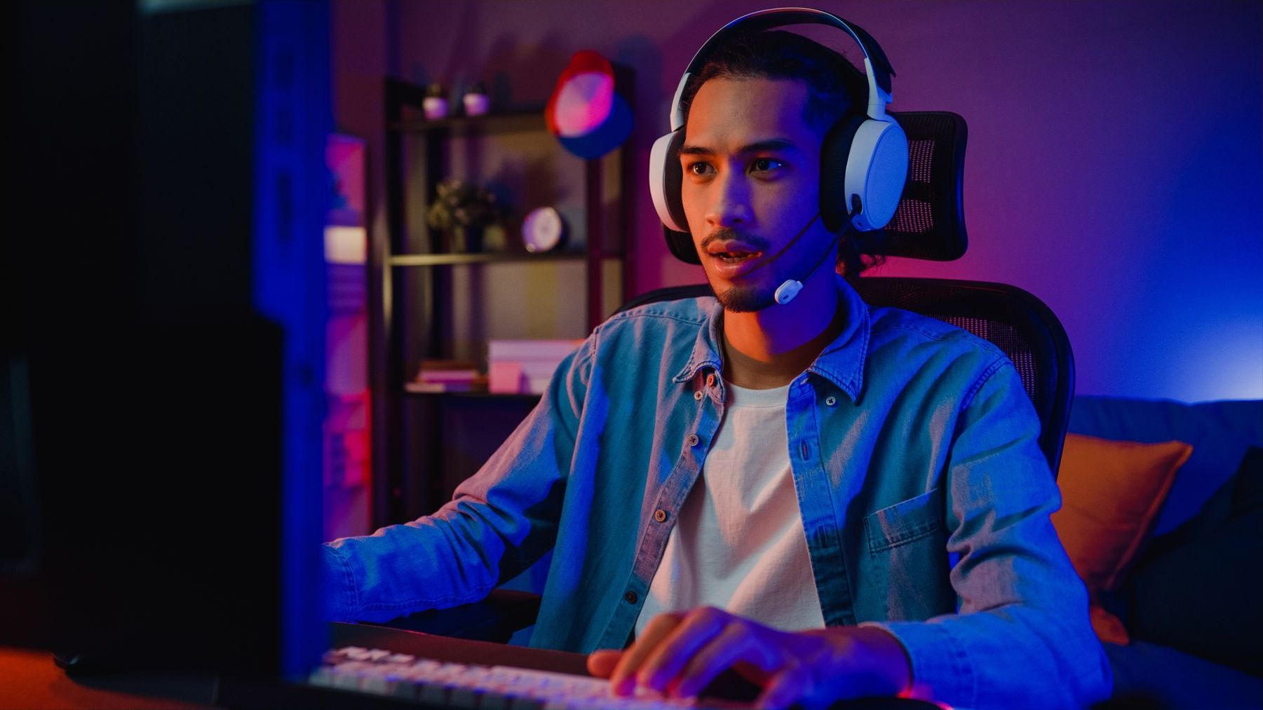 Man sitting in an Ergohuman gaming chair playing at his PC. He is wearing white headphones with a mouthpiece and looks intensely at the screen. The lighting is low and moody with a pink and blue glow. 