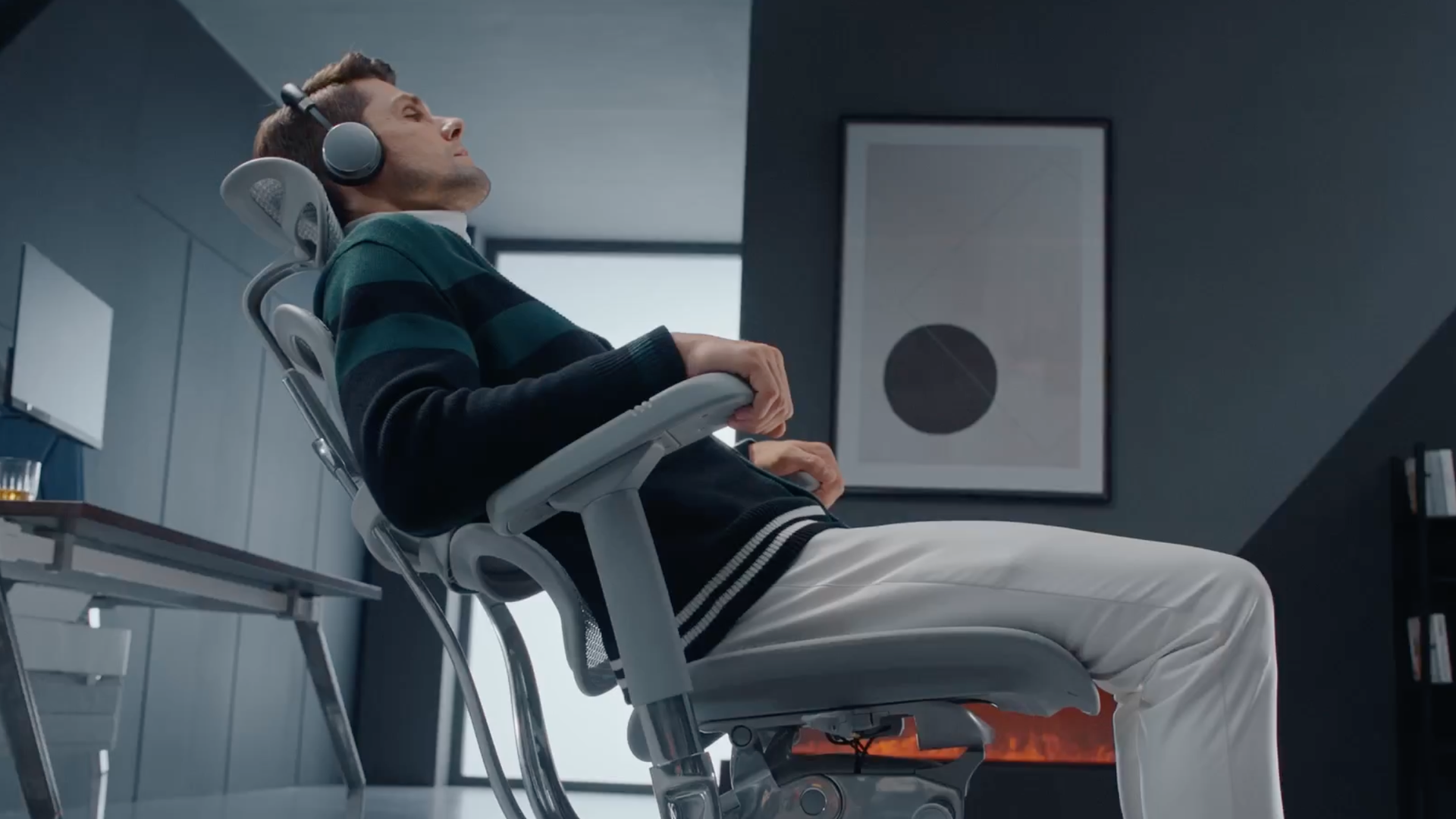 A man is relaxing in recline mode in his Ergohuman Elite. It's a grey frame with grey mesh. The man is wearing a dark sweater and white jeans. He is wearing headphones and relaxing. The room he's in is dark grey with natural lighting. There is a fire on in the background. 