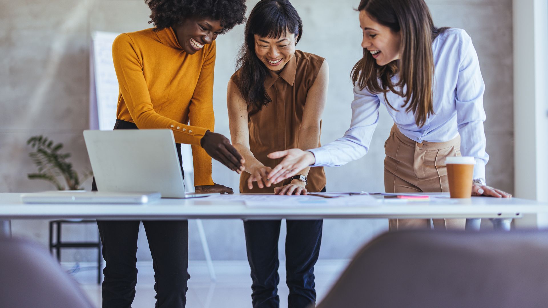 Three women stand at a desk while working. They are smiling and gesturing to the paper and laptop on the desk. 