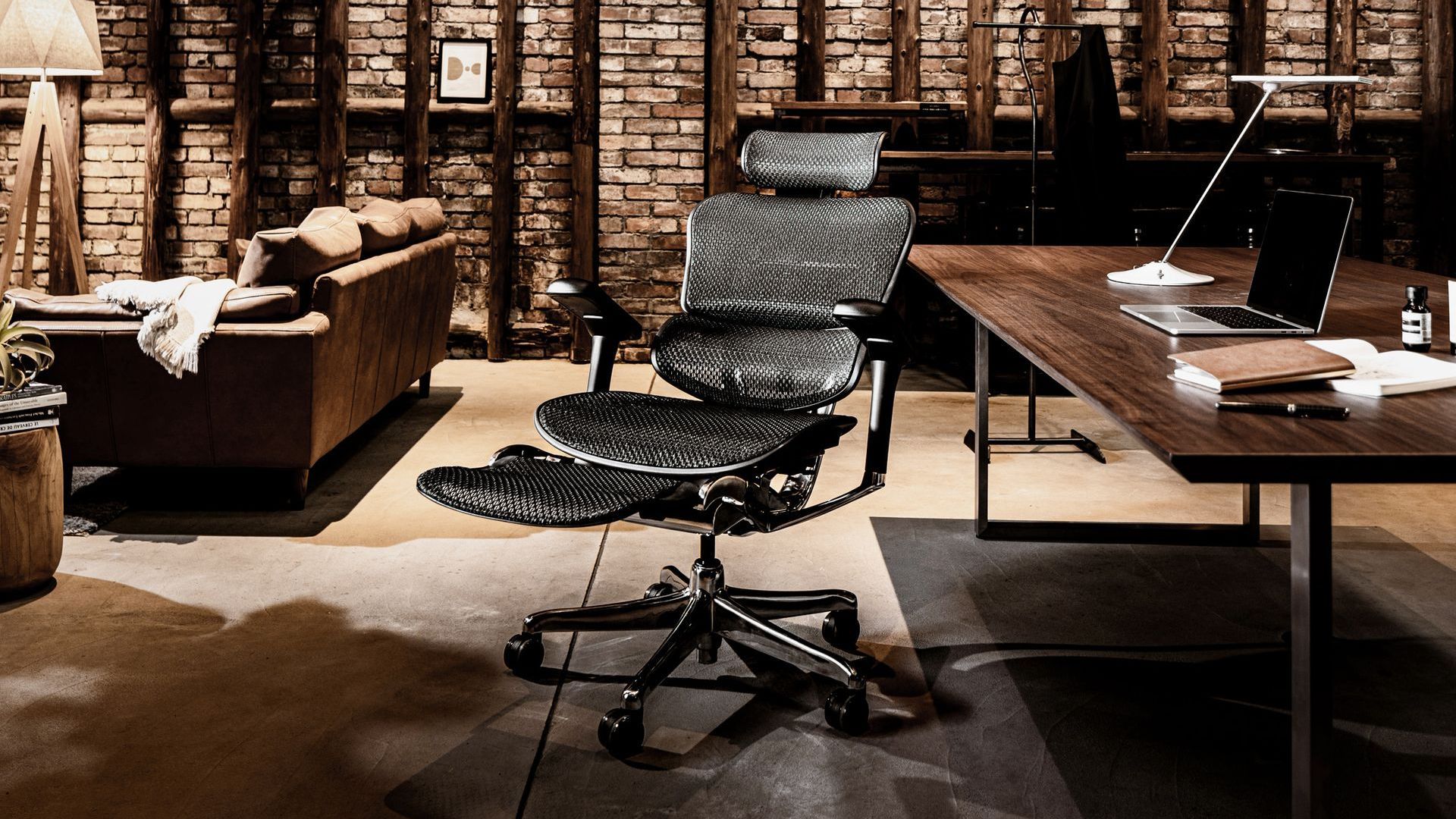 A modern lounge style room with brick walls and leather sofa has a black mesh office chair with a legrest in a reclined position sitting beside a desk.