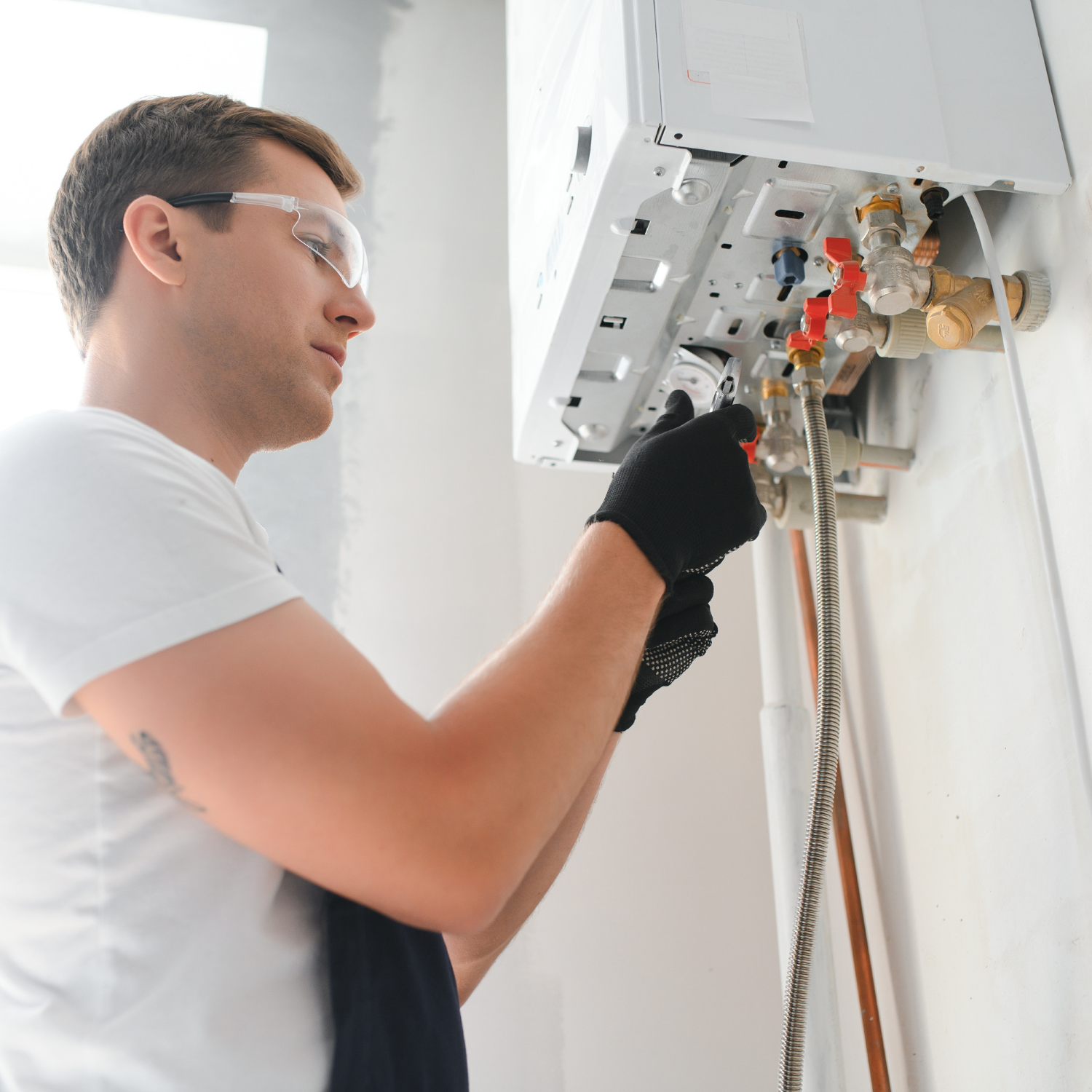 Man in safety glasses and gloves fixing a white wall-mounted boiler, in a room with white walls.