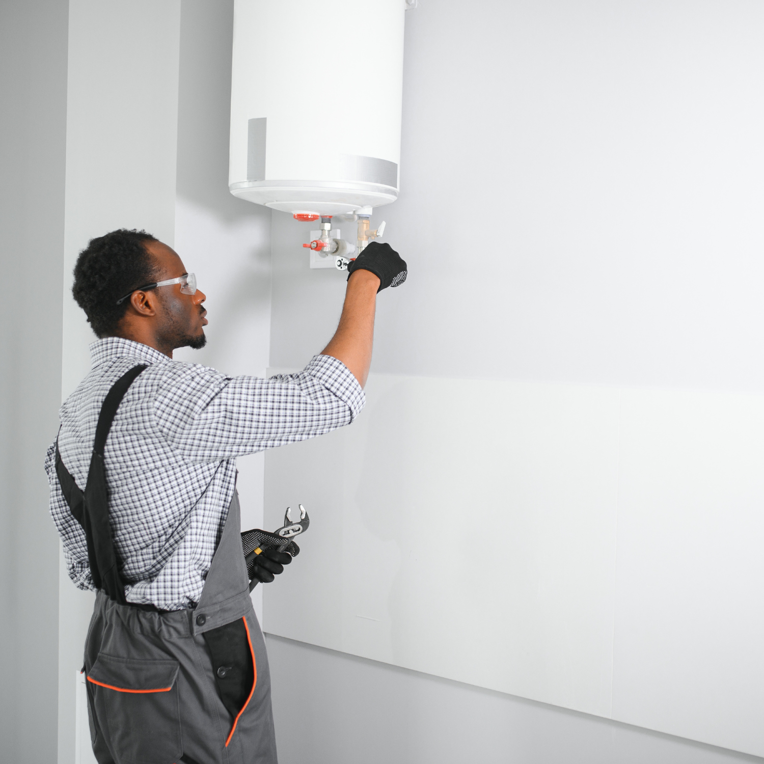A person in work clothes, safety glasses, and gloves working on a white water heater mounted on a wall.