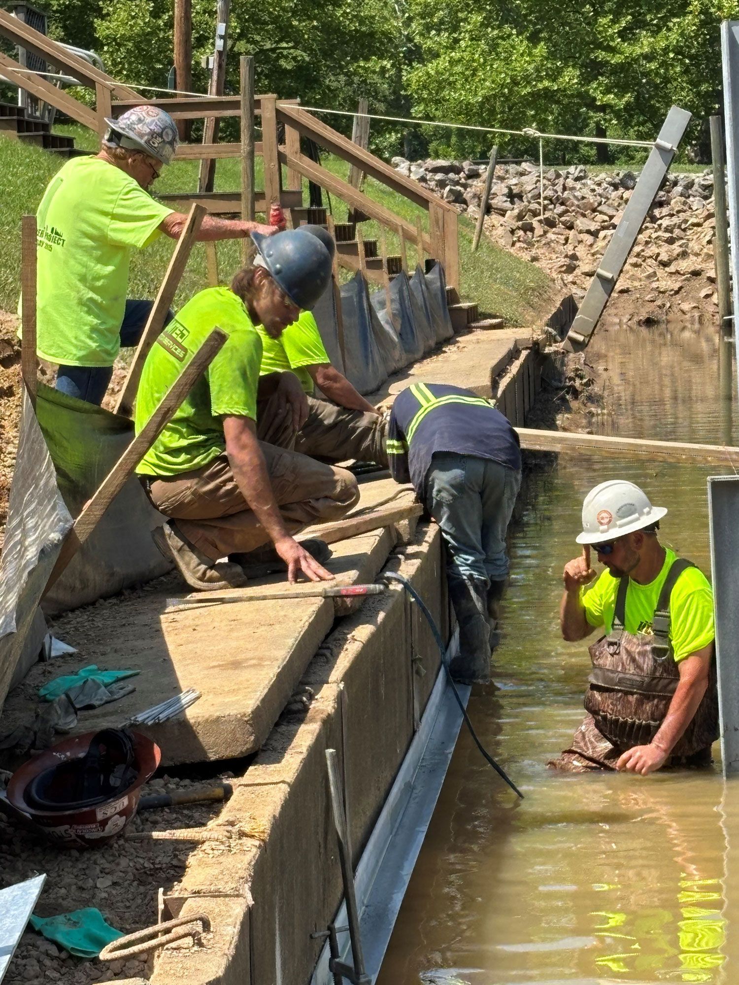 Construction workers installing barrier in a shallow, murky water. One worker standing, others kneeling or in water.