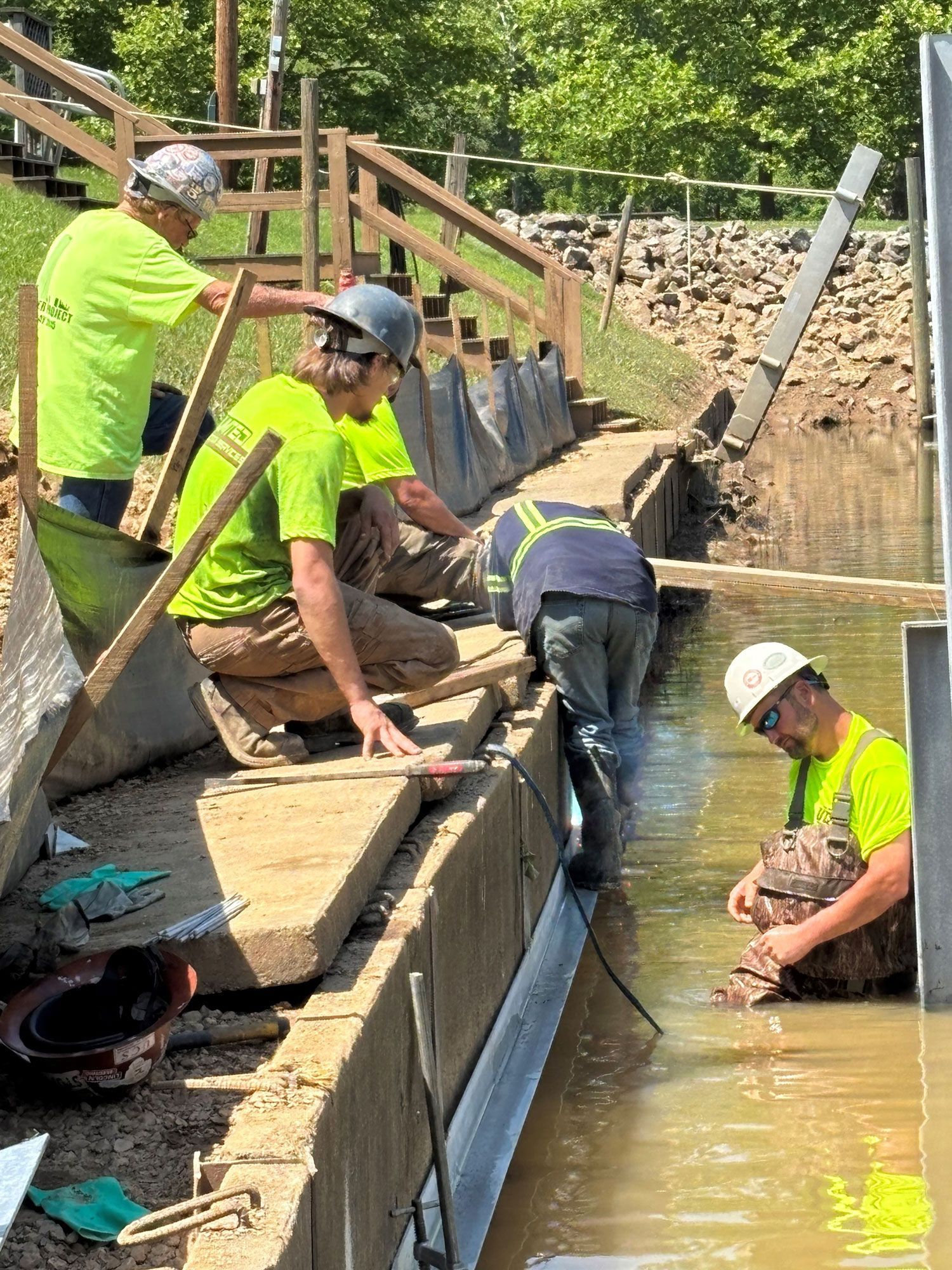 Construction workers building near water; green shirts, hard hats. Sunny outdoor setting, river.