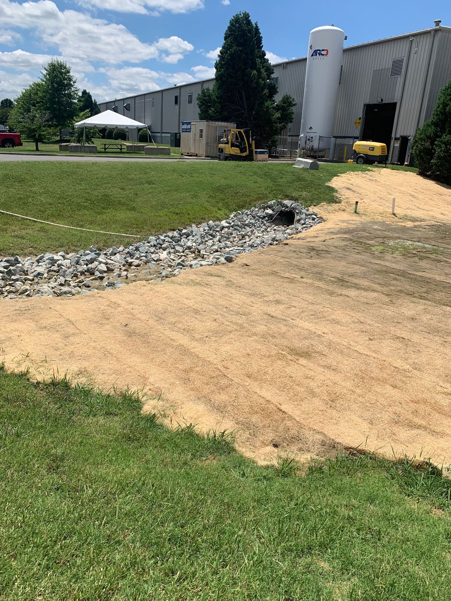 Grassy area with gravel and tan mulch, industrial building, trees, blue sky.