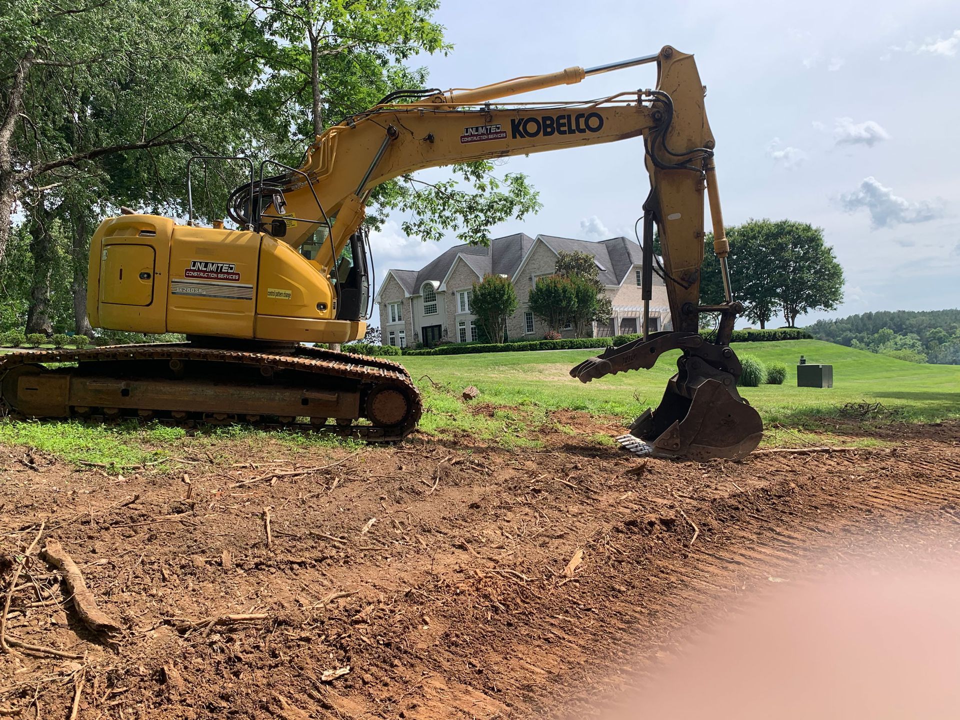 Yellow excavator on a dirt field, digging with a house in the background.