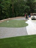 A putting green with flag, surrounded by gray stone and green grass, in a backyard with trees.