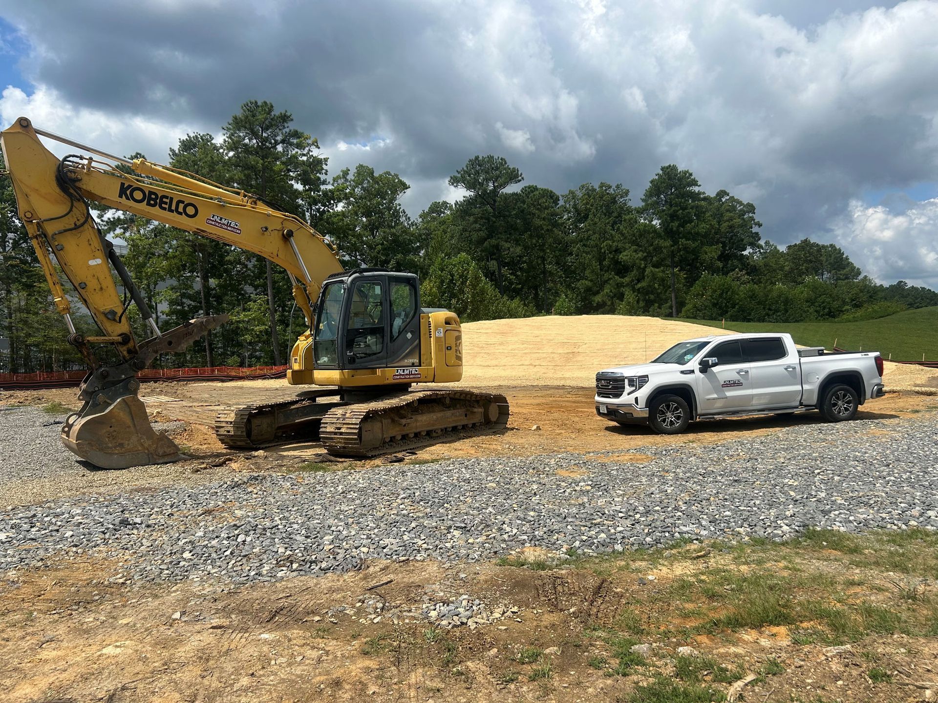 Yellow excavator and white truck on gravel at a construction site, trees in background.