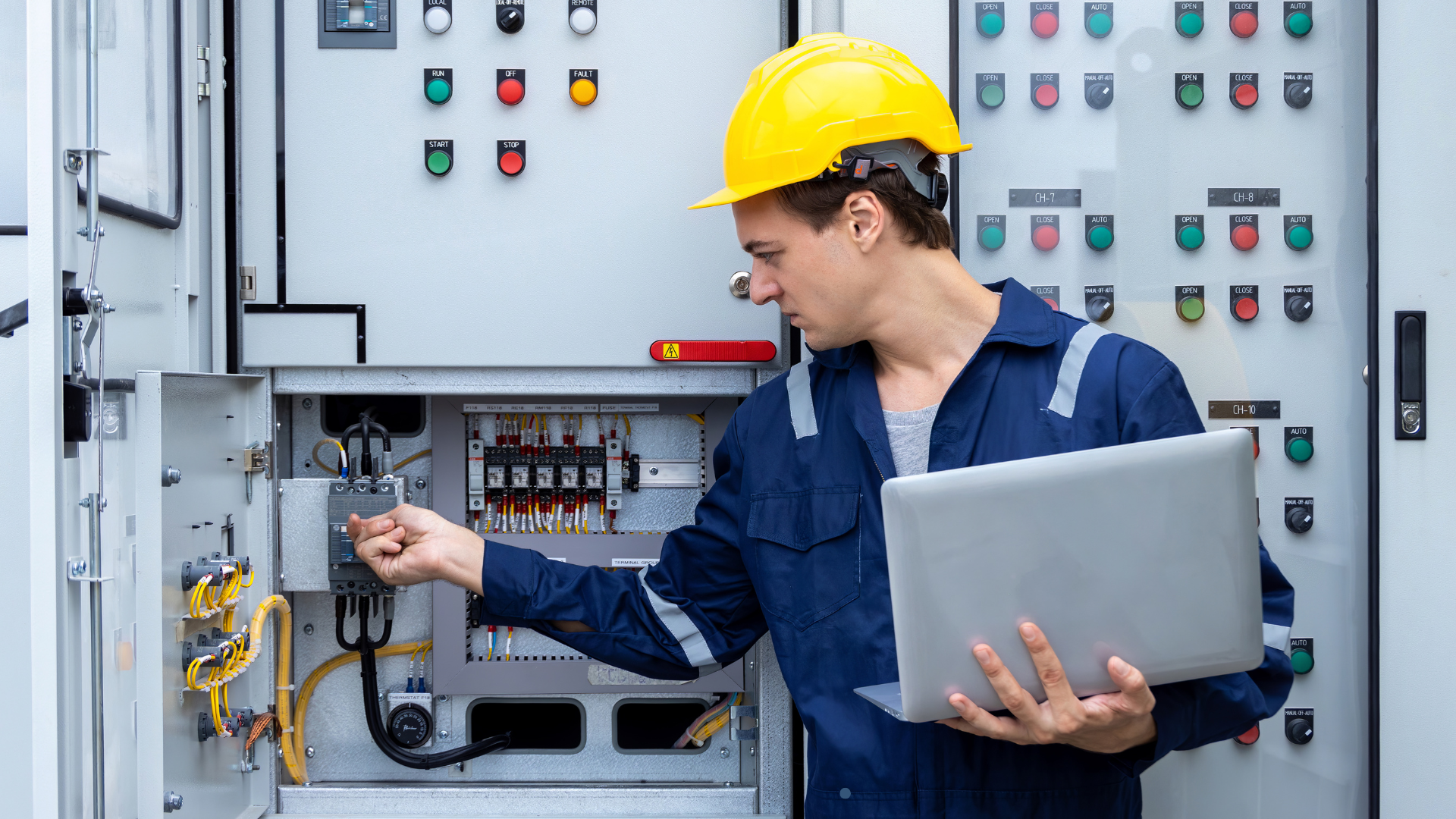 An engineer wearing a yellow hard hat operates electrical systems while holding a laptop and adjusti