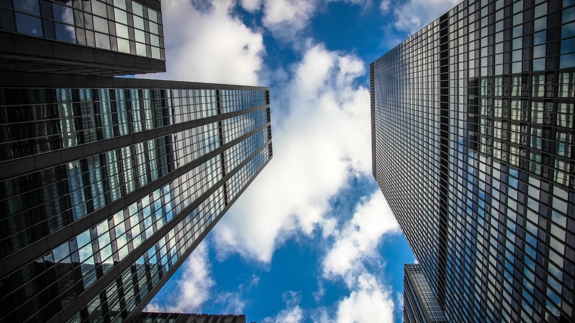 A low-angle view of modern high-rise buildings, reflecting the sky and clouds in their glass facades