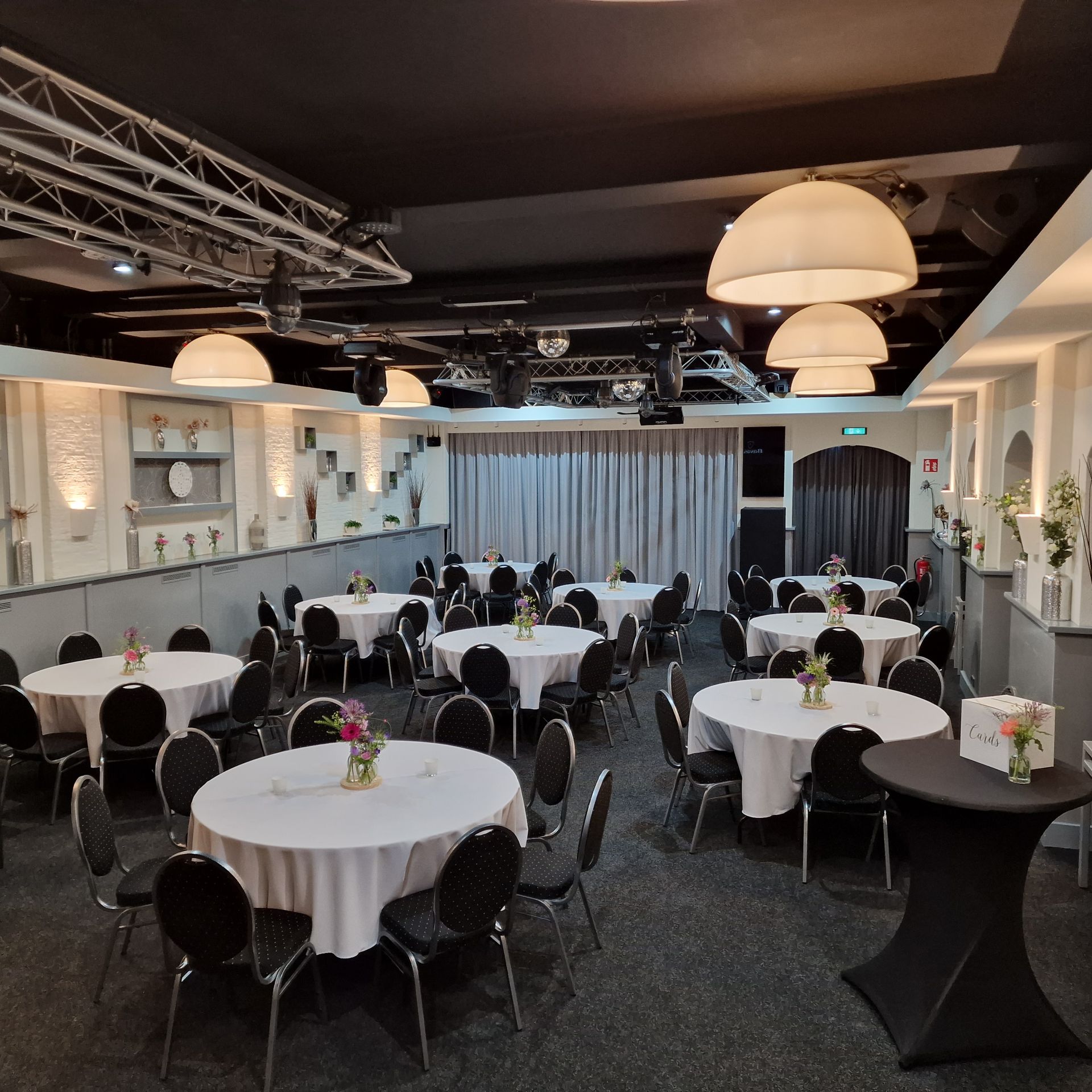 Evenementenruimte met ronde tafels gedekt met witte tafelkleden en zwarte stoelen, klaar voor de gasten.