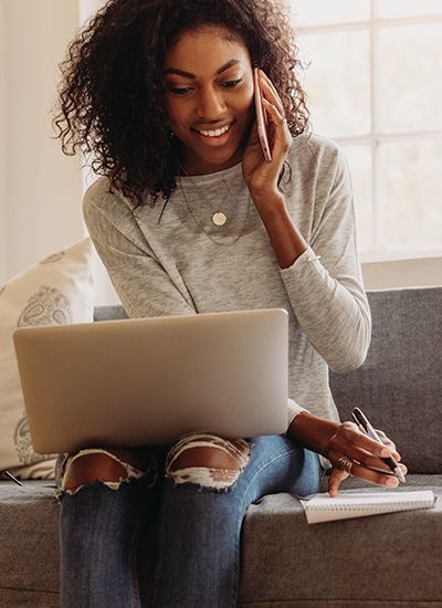 A woman is sitting on a couch using a laptop and talking on a cell phone.