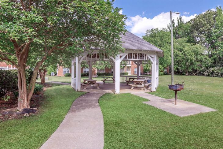 Gazebo with picnic tables in a grassy park, a pathway leads to it, barbecue grill nearby, trees surround it.