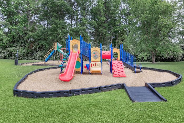 Playground with a red slide, blue structure, surrounded by tan ground and grass.