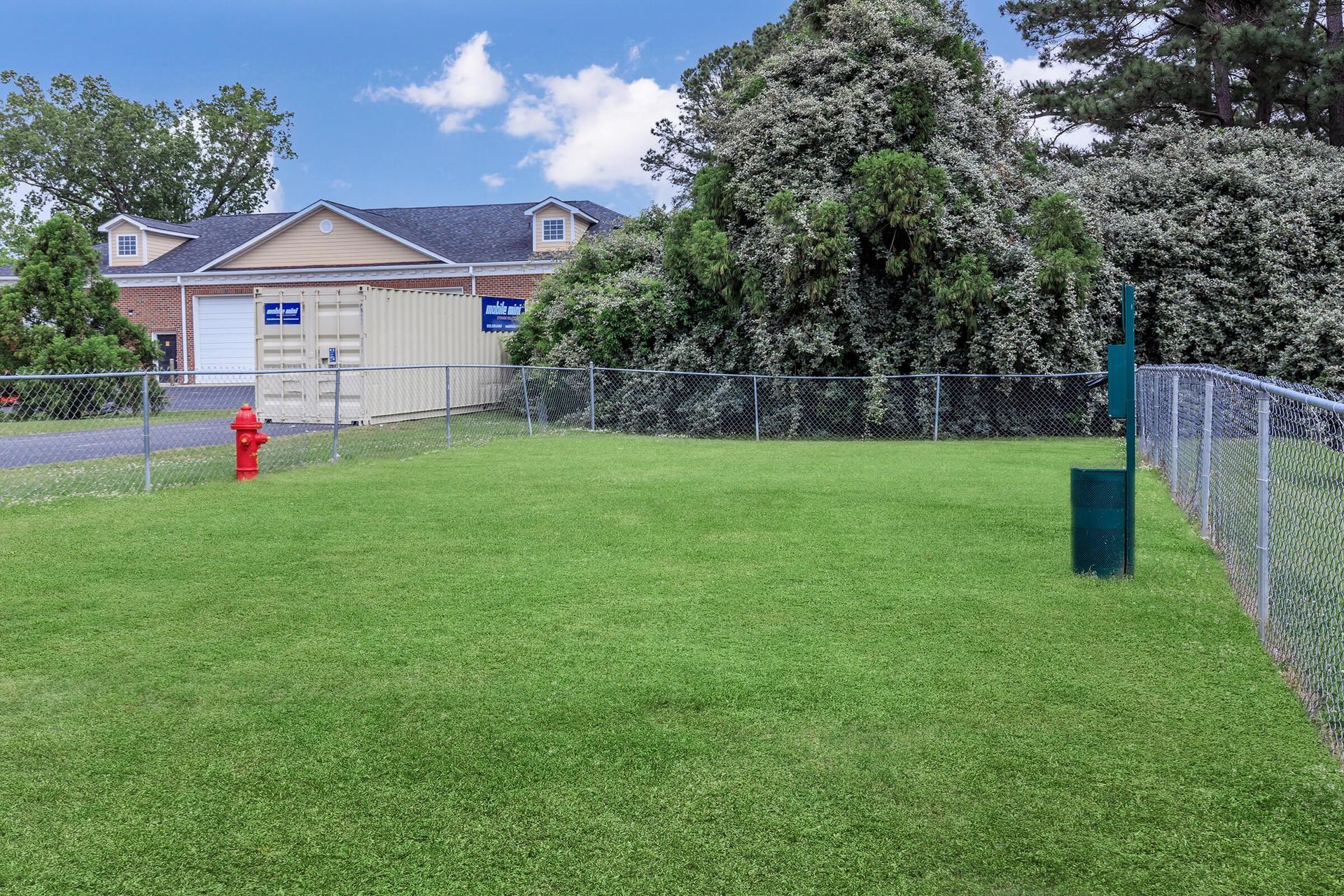 Grassy fenced dog park with a red fire hydrant and waste bag dispenser. A building and trees are in the background.