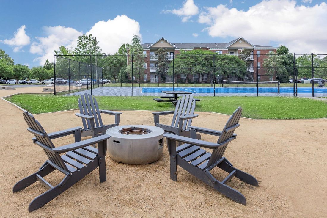 Four gray Adirondack chairs around a fire pit, with a blue recreational court and apartment building in the background.
