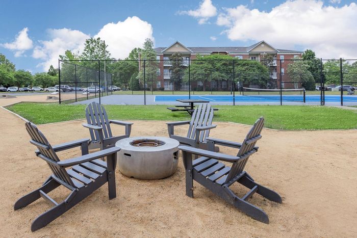 Four gray Adirondack chairs around a fire pit, with a blue recreational court and apartment building in the background.