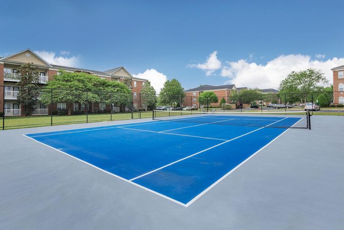 Blue tennis court outdoors with apartment buildings in the background on a sunny day.