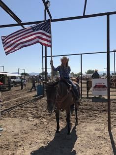 A person in a cowboy hat rides a horse in an outdoor arena while holding an American flag.