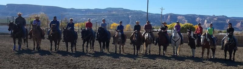 A group of people on horseback lined up in a dirt arena against a backdrop of mountains and trees.