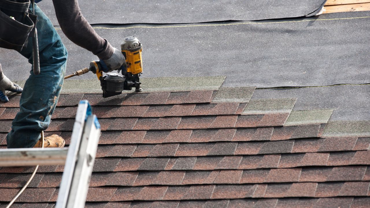 A worker uses a nail gun to install brown asphalt shingles on a sloped roof.