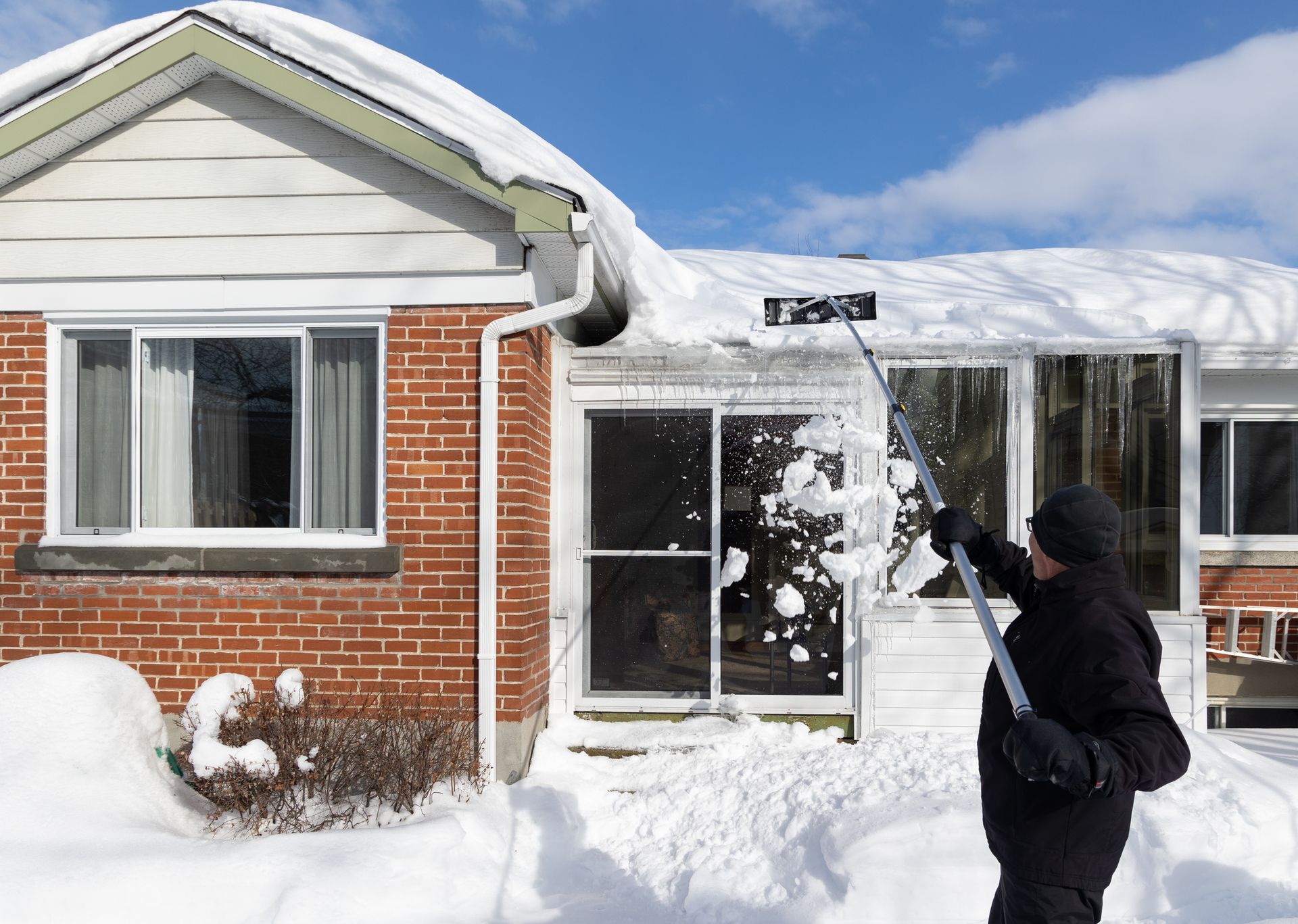 Person uses a snow rake to remove snow from a roof on a sunny day.