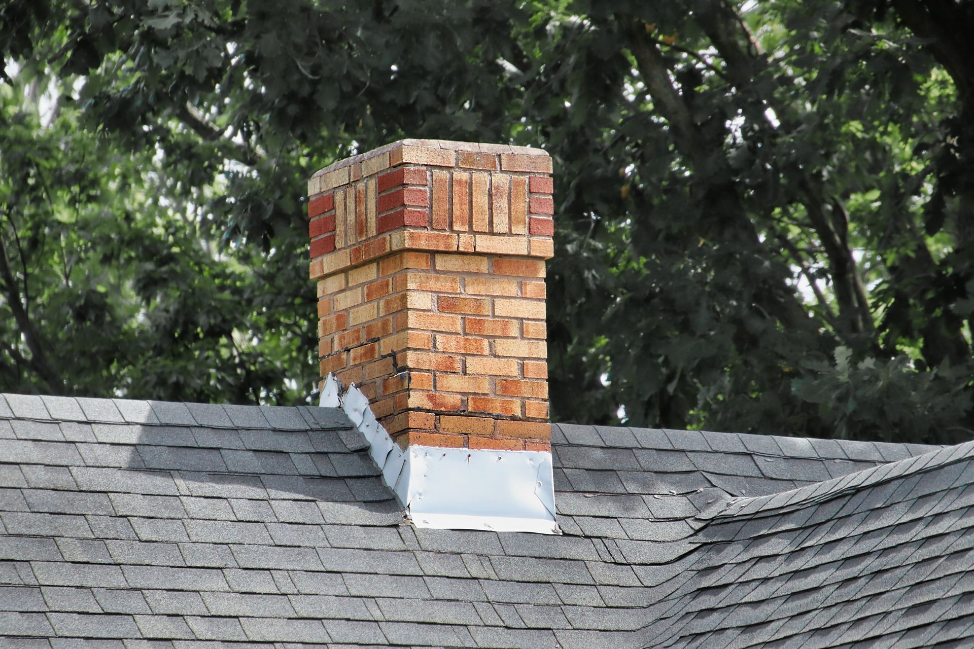 Brick chimney on a gray shingled roof, with trees in the background.