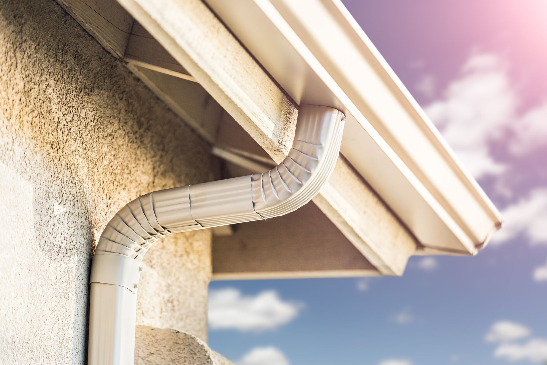 White gutter system on a beige stucco house under a blue sky with pink hues.