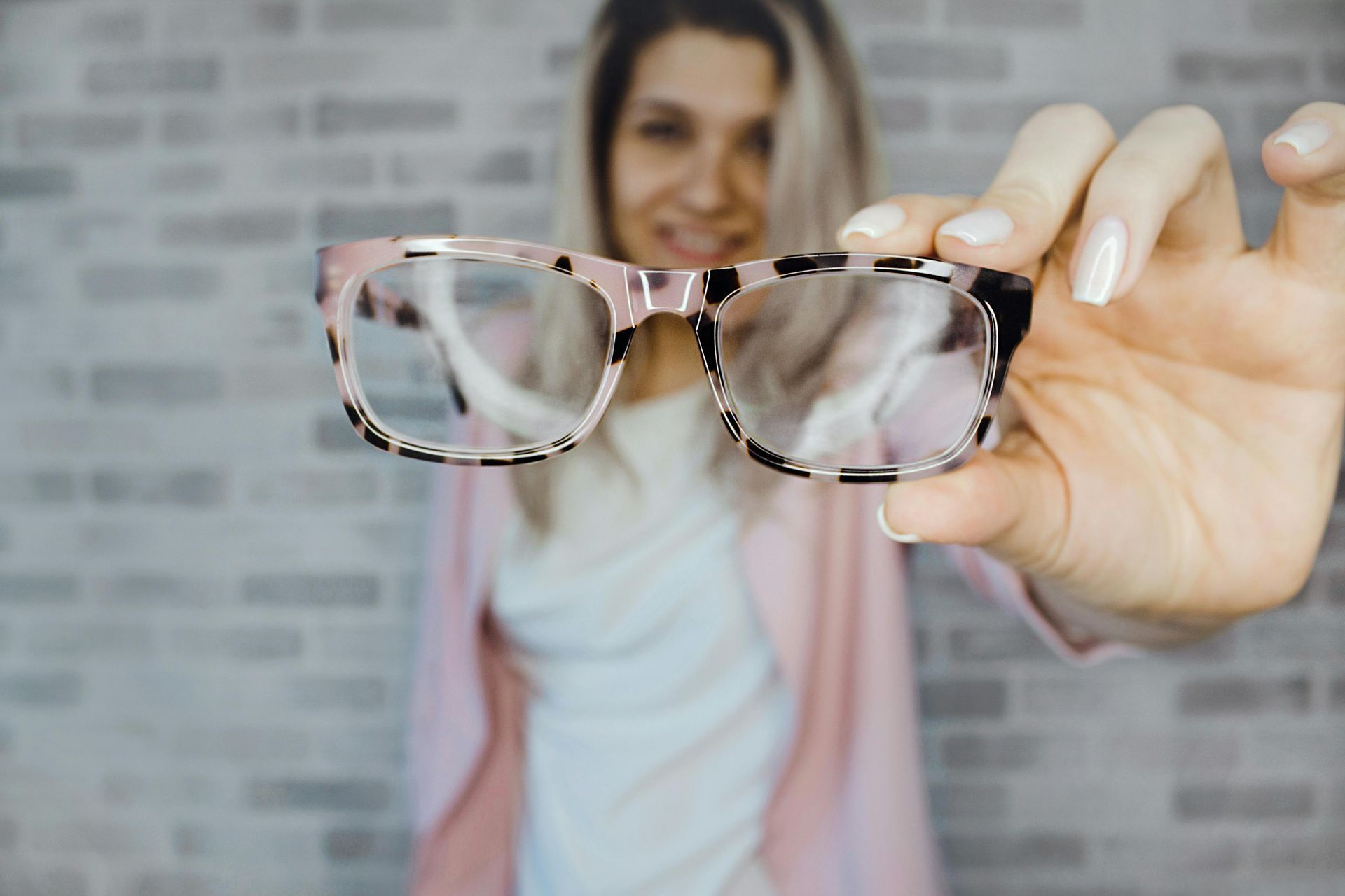 Woman holding up glasses with pink and leopard-print frames against a brick wall.
